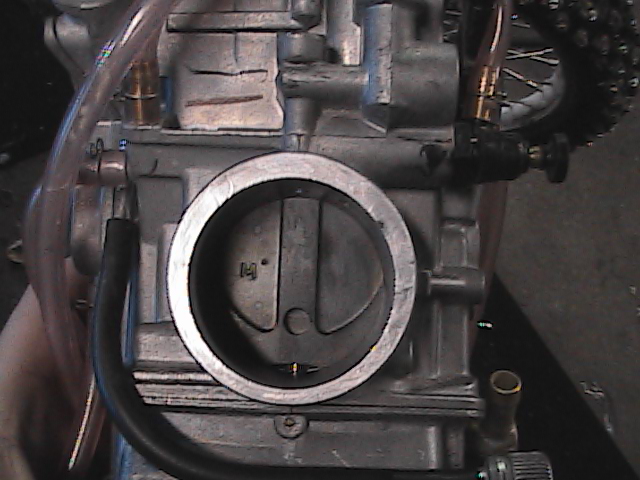 Motorcycle carburetor with a circular intake and visible throttle plate. A chain and spokes are visible in the background