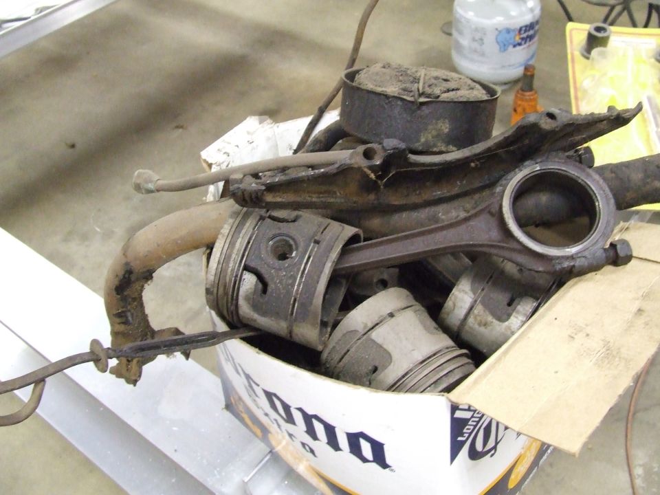 Corona box full of dirty used pistons, a connecting rod, and rusty engine parts on a shop table