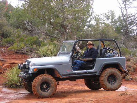 1983 Jeep CJ7 - A man in a hat drives a silver 1983 Jeep CJ7 through a rocky, desert landscape.