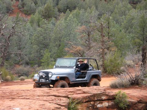 1983 Jeep CJ7 - A driver waves from a gray 1983 Jeep CJ7 navigating a rocky trail in a wooded, arid landscape.