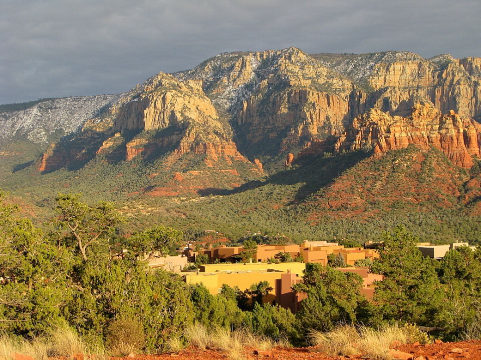 Mountain landscape with red rock formations, scattered green trees, and a cluster of tan buildings in the mid-ground. Snow is visible on the upper slopes of the mountains