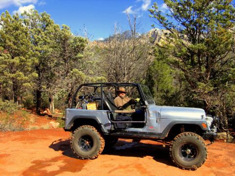1983 Jeep CJ7 - A man drives a silver Jeep Wrangler through a wooded, mountainous area on a sunny day.