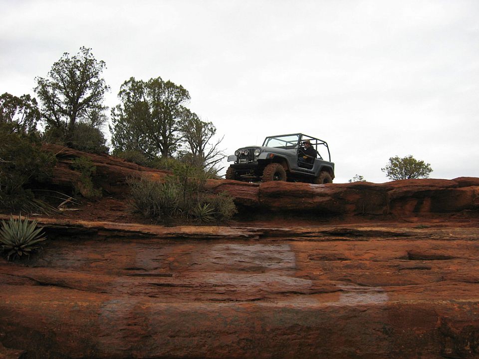 Jeep Wrangler driving on a rocky, off-road trail with trees and a cloudy sky