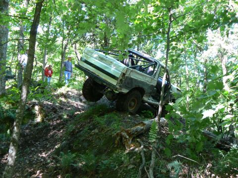 1995 Ford Bronco - This modified SUV is navigating a steep, wooded incline with significant body damage and a visible roll cage.