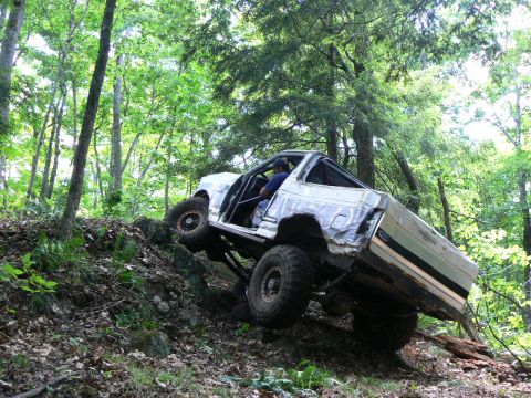 1995 Ford Bronco - Modified off-road SUV navigates a challenging, rocky incline in a dense forest.