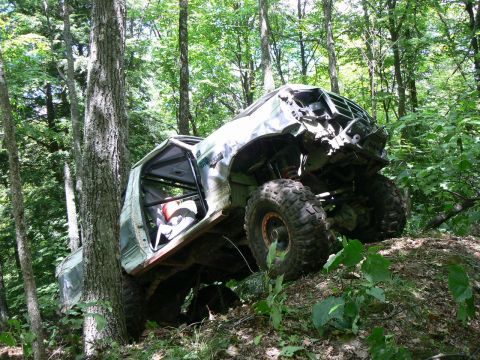 1995 Ford Bronco - Modified 1995 Ford Bronco with significant front-end damage, stuck against a tree in a wooded area.