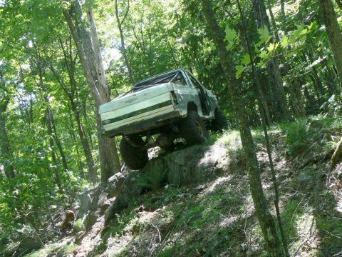1995 Ford Bronco - A modified off-road vehicle navigates a rocky incline in a dense forest setting.