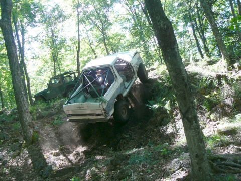 1995 Ford Bronco - A white off-road SUV, modified with a roll cage, navigates a challenging incline in a wooded area, with another vehicle in the background.