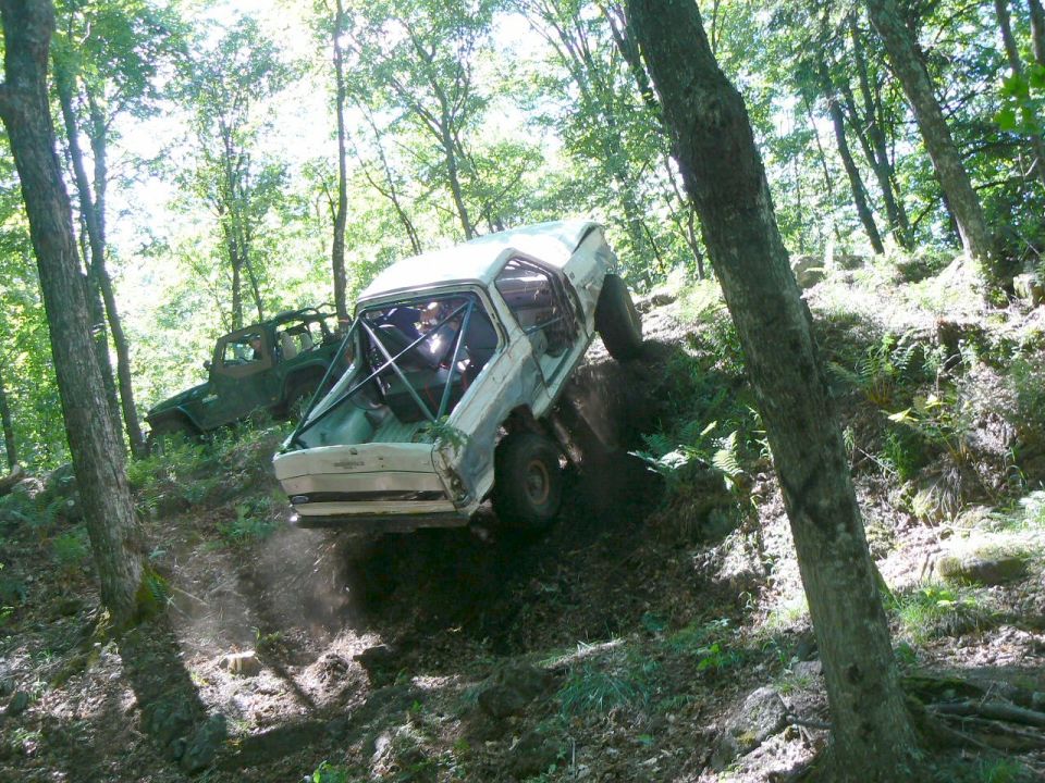 White off-road SUV with roll cage climbing a steep, wooded hill. Another green SUV is visible behind it