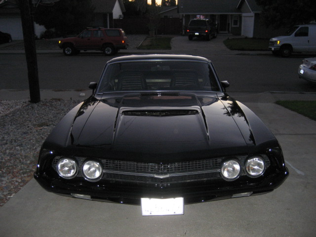 Black classic Ford with dual headlights and hood scoop, front view on driveway at dusk