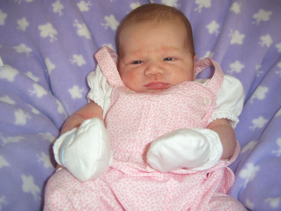 Newborn baby in pink overalls and white mittens, lying on a purple blanket with white stars