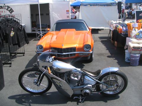 1986 Pontiac Fiero - An orange classic car and a custom motorcycle are parked outdoors near vendor tents.