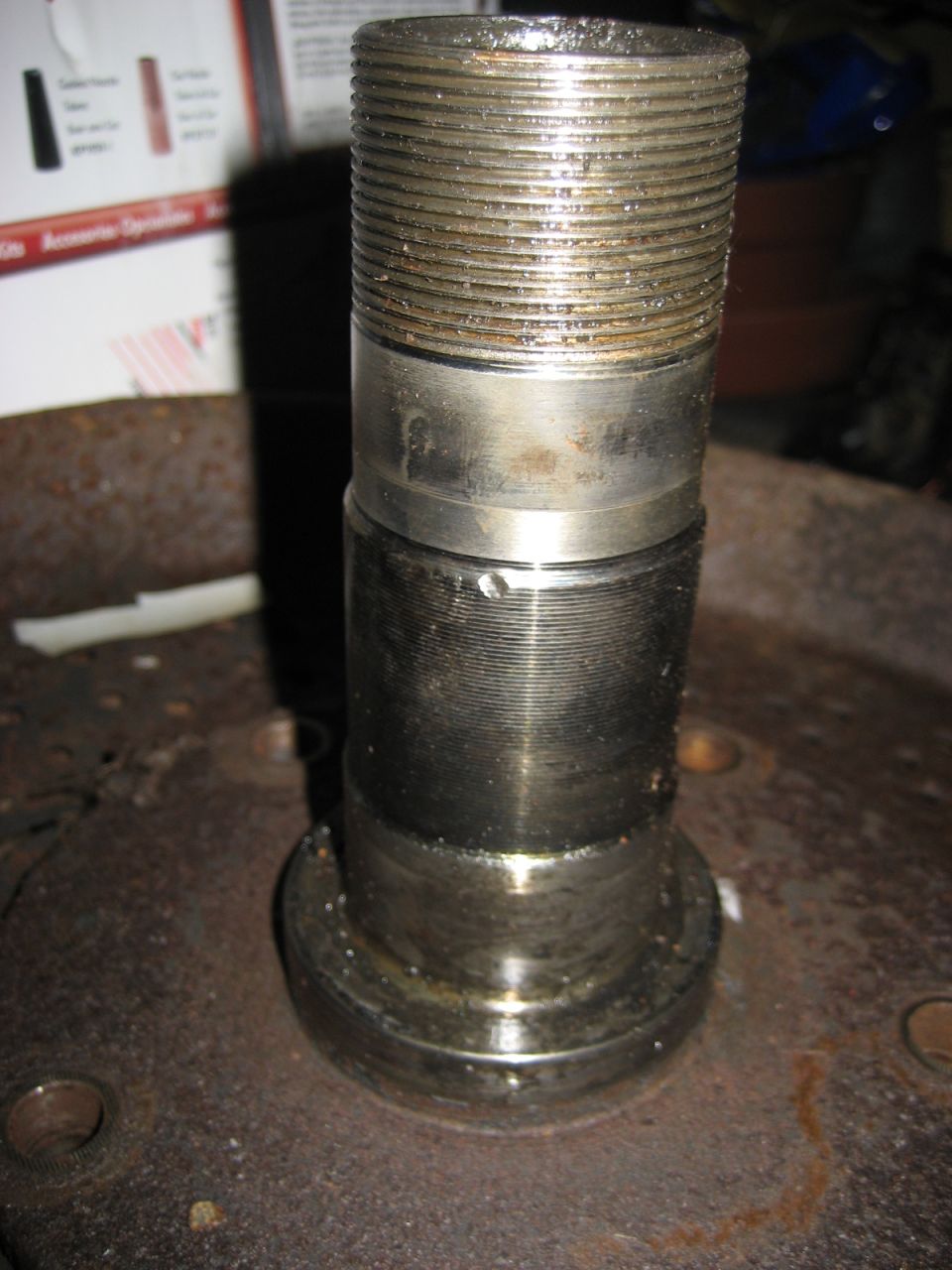 Steel front axle spindle standing on a rusty bench showing threads and bearing journals