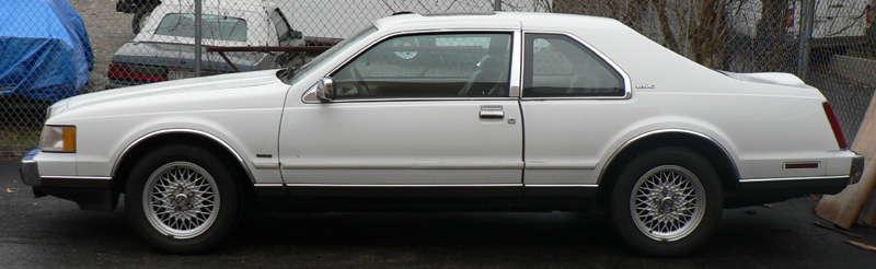 White Lincoln Mark VII coupe in side profile with mesh alloy wheels, parked by a chain-link fence