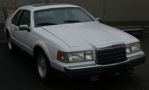 1990 Lincoln Mark VII - A white Lincoln Mark VII is shown from a three-quarter front angle, parked outdoors with a fence in the background.