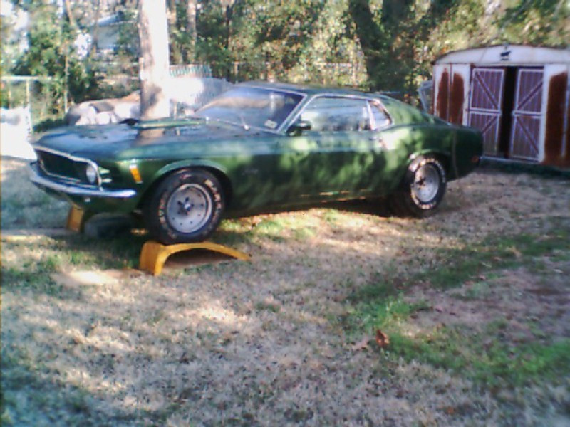 Green Ford Mustang fastback on yellow ramps in a grassy yard, front 3/4 view, shed behind