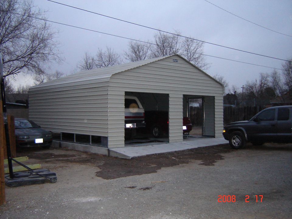 Metal garage with two bays, one occupied by a white SUV and a dark car. Another red car is visible in the second bay. A dark SUV is parked outside to the right