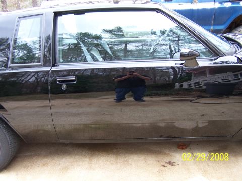 1979 Ford F-150 - A black car is seen from the side, with a reflection of a person and trees. Another vehicle is partially visible.