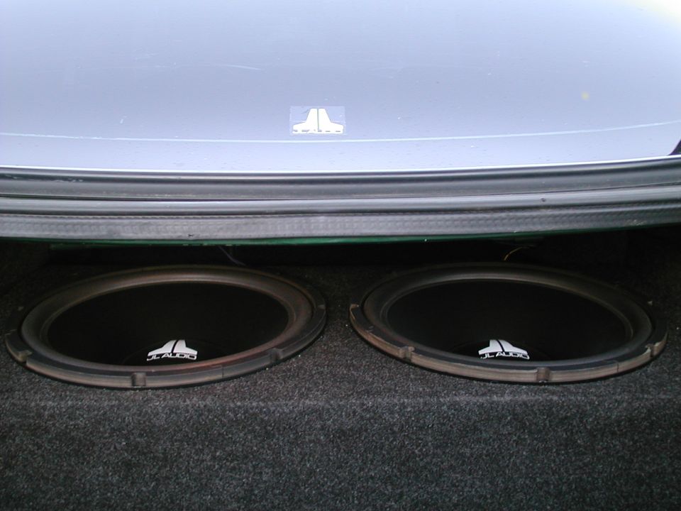 Two JL Audio subwoofers mounted under a rear deck in a carpeted trunk with JL emblem above