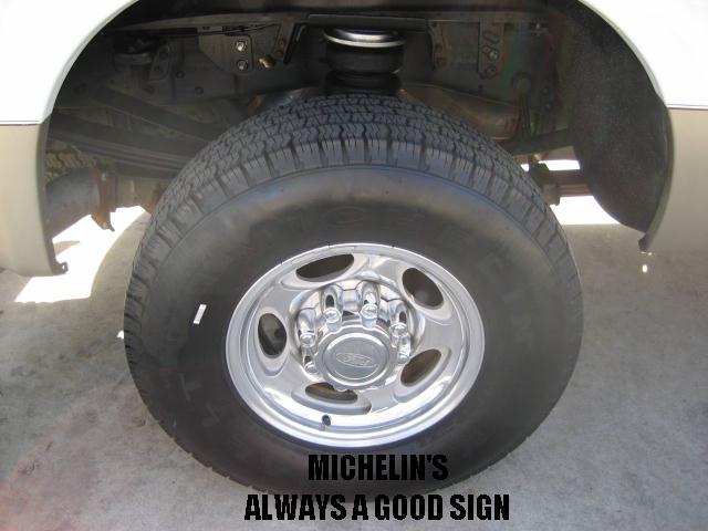 Close-up of a truck's rear wheel and suspension components. The wheel has a chrome finish with five large oval cutouts