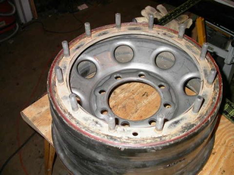1990 Ford Bronco - A silver 8-lug wheel sitting face-up on plywood with its beadlock ring removed, exposing a ring of threaded studs standing up around the re…