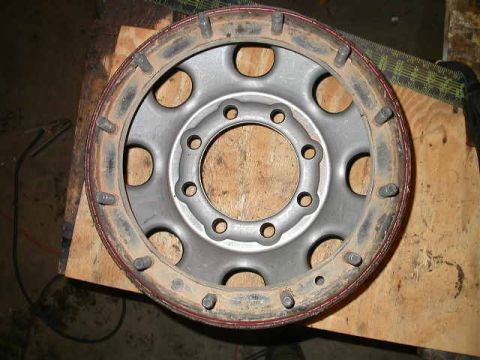 1990 Ford Bronco - Overhead view of a silver 8-lug beadlock wheel lying face-up on a plywood sheet, its outer lock ring held by a full circle of studs and nut…