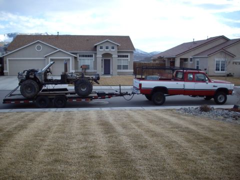 1982 Toyota 4Runner - A red and white pickup truck tows a trailer carrying a bare off-road vehicle frame down a suburban street.