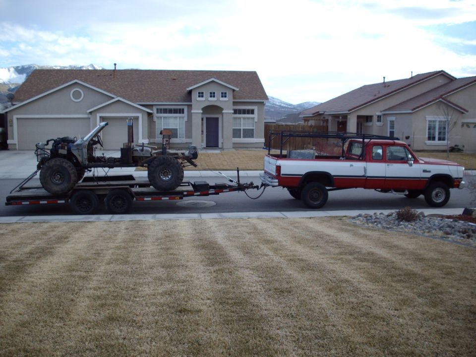 Pickup truck towing a trailer with an off-road vehicle chassis. The chassis is stripped down with large tires