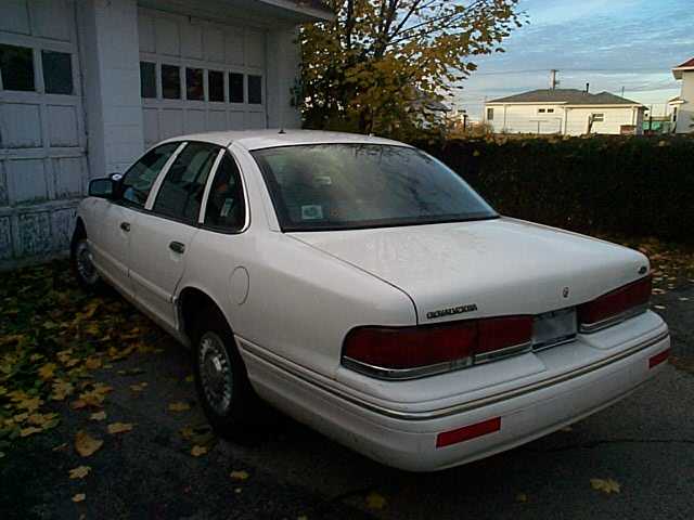 White full-size sedan viewed from rear driver's side, parked by a garage with fallen leaves.
