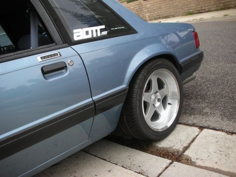 1990 Ford Mustang - A blue 1990 Ford Mustang is parked on a street, with a close-up on its rear wheel and door panel.