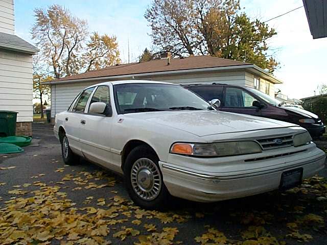 White Ford Crown Victoria with wire-look hubcaps parked on leaves by a garage and another car