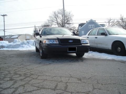2000 Ford Crown Victoria - A black 2000 Ford Crown Victoria is parked next to a white vehicle on a cracked asphalt lot with snow piles.