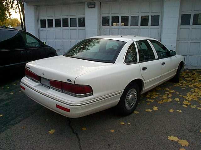 White Ford Crown Victoria sedan, rear passenger-side view, parked on leafy driveway by white garage doors