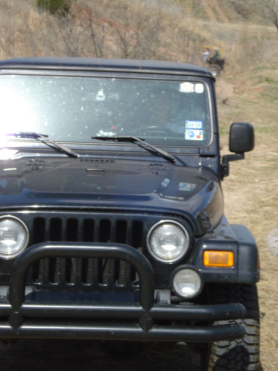 Dark blue Jeep Wrangler front grille with bull bar, dusty windshield, and a distant ATV on a hill