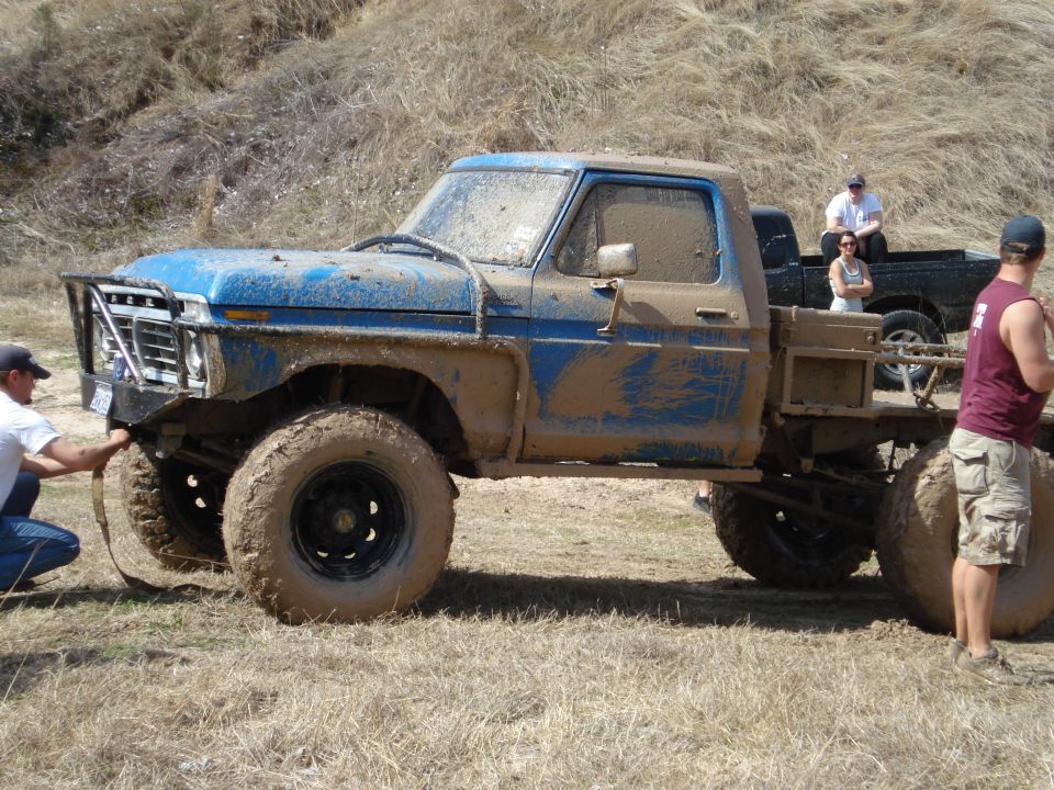 Mud-covered blue pickup truck with large tires and a custom bumper, parked on dry grass. Another pickup truck is visible behind it