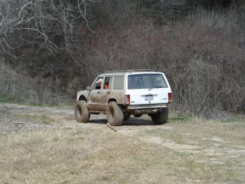 2008 International School Bus - A white Jeep Cherokee, modified for off-roading, navigates a muddy trail through a wooded area.
