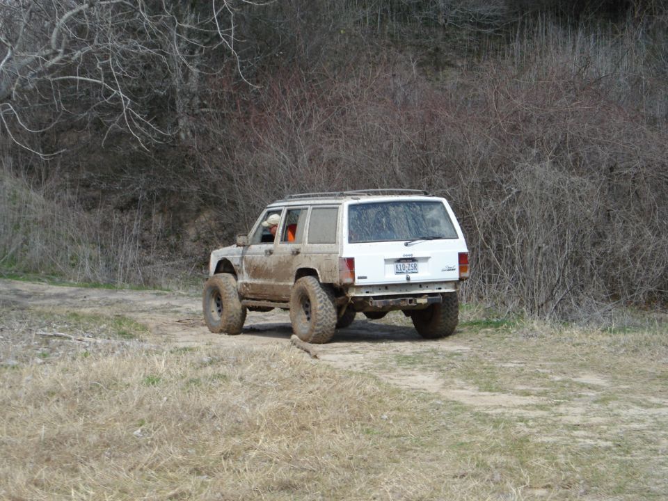 White Jeep Cherokee with big tires driving on a dirt path, covered in mud. Bare trees and brush in background