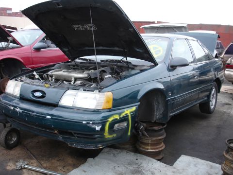 1993 Ford Taurus - A dark green 1993 Ford Taurus SHO sedan sits in a junkyard with its hood open and front wheels removed.