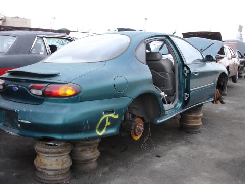 1993 Ford Taurus - Junkyard Ford Taurus, missing wheels and doors open, sits on tires with other vehicles nearby.