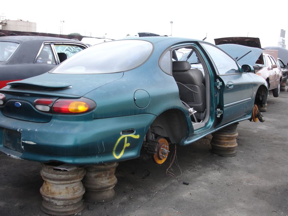 Green Ford Taurus sedan missing wheels, doors open, sitting on tires in junkyard. Another car is visible behind it