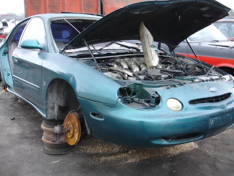 1993 Ford Taurus - A teal sedan sits in a junkyard with its hood open, revealing the engine compartment and missing front passenger wheel.