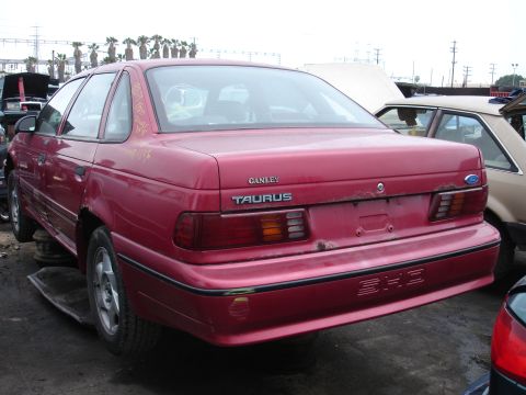 1993 Ford Taurus - A red Ford Taurus sits in a junkyard, showing its rear end and taillights.