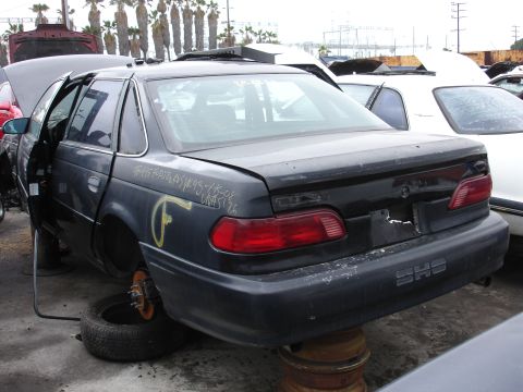 1993 Ford Taurus - A dark sedan, possibly a 1995 Ford Taurus SHO, sits in a junkyard with its wheels removed and doors ajar.