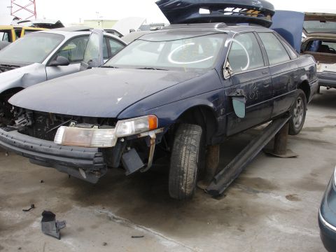 1993 Ford Taurus - A damaged blue sedan is elevated on wooden blocks in a junkyard setting, with other vehicles visible.