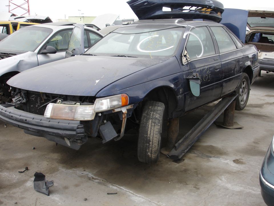 Blue sedan on blocks, front end damaged, missing grille and headlight. Another car is to the left