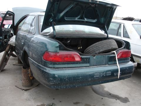 1993 Ford Taurus - Junkyard scene with a 1993 Ford Taurus, showing its rear and open trunk with a spare tire.