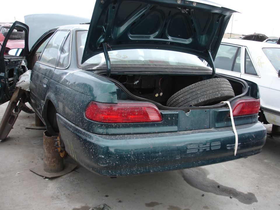 1993 Ford Taurus rear view in junkyard, trunk and hood open, spare tire visible in trunk