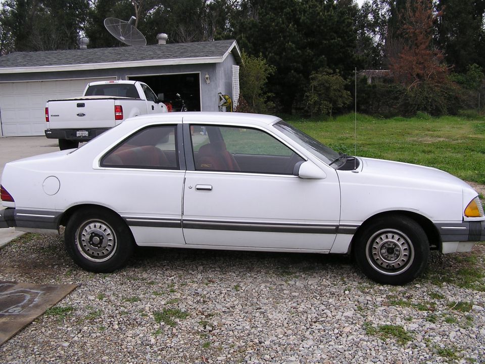 White 1980s Ford Tempo two-door coupe parked on a gravel driveway with a pickup in the background