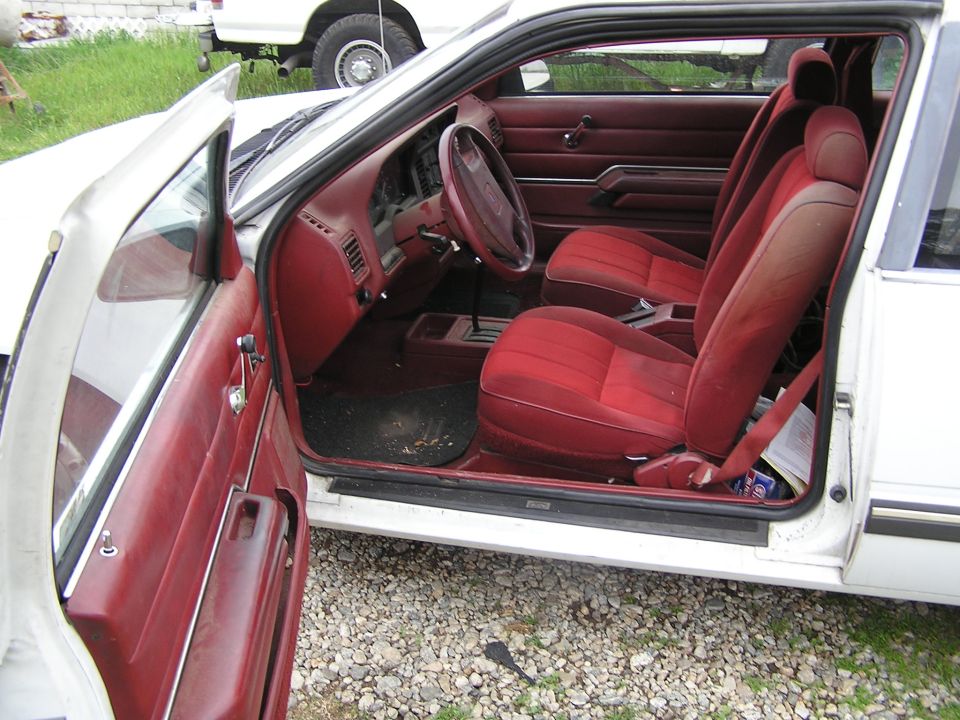 Interior of white coupe with red cloth seats and dash, door open, on a gravel lot