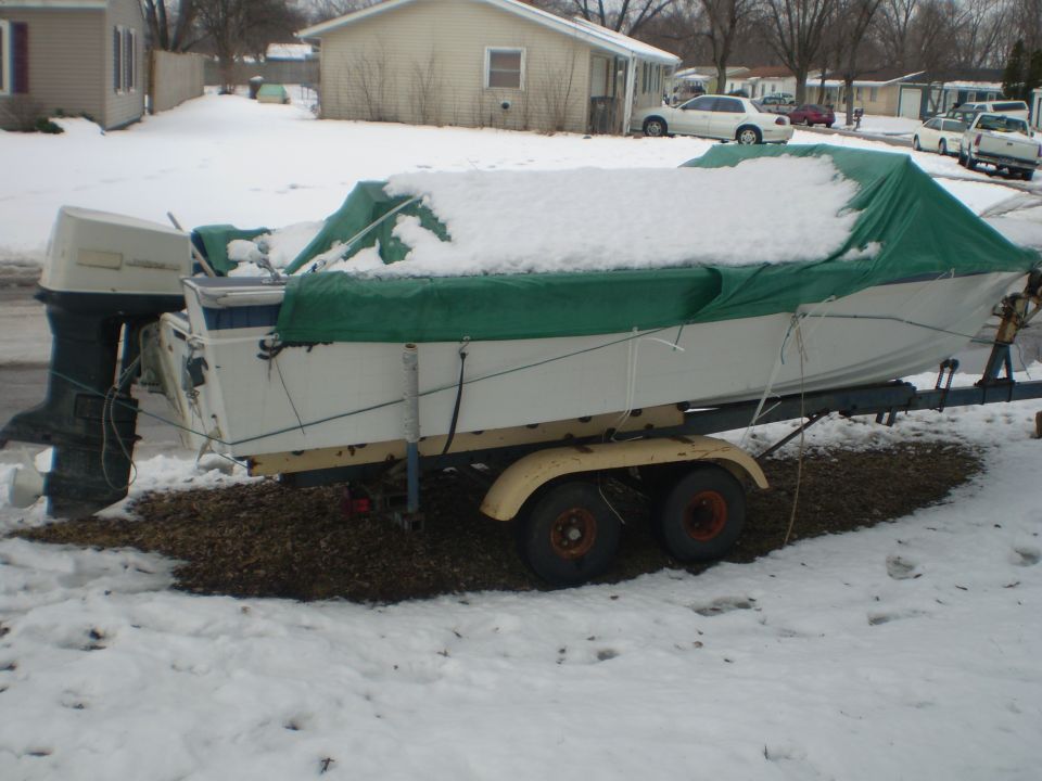 White boat with green cover and outboard motor on a tandem-axle trailer in a snowy yard
